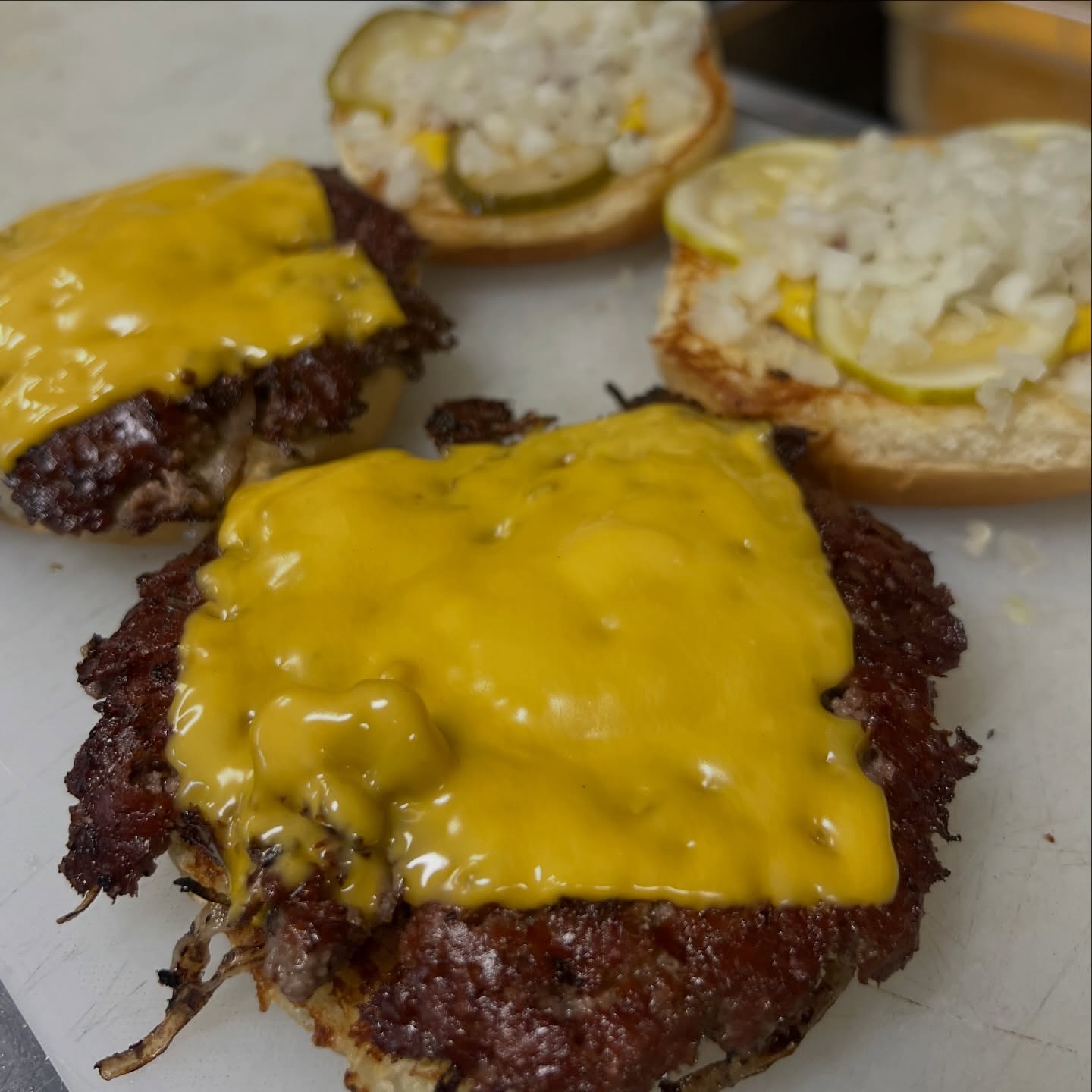 Smashed beef patty with melted American cheese, beside open buns dressed with pickles and onions